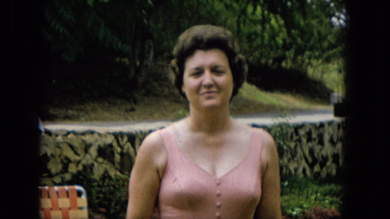 The woman faces the camera in the garden, wearing a pink bathing suit, with trees, a low stone wall, and a striped folding chair partly visible.