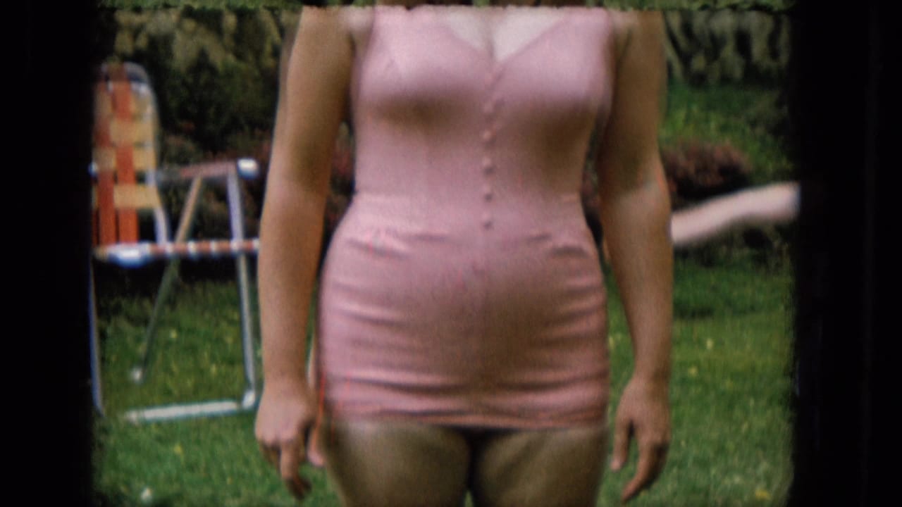 Fuller 1965 home-movie view of the woman standing on a lawn in a pink bathing suit, with garden greenery and a striped chair in the background.