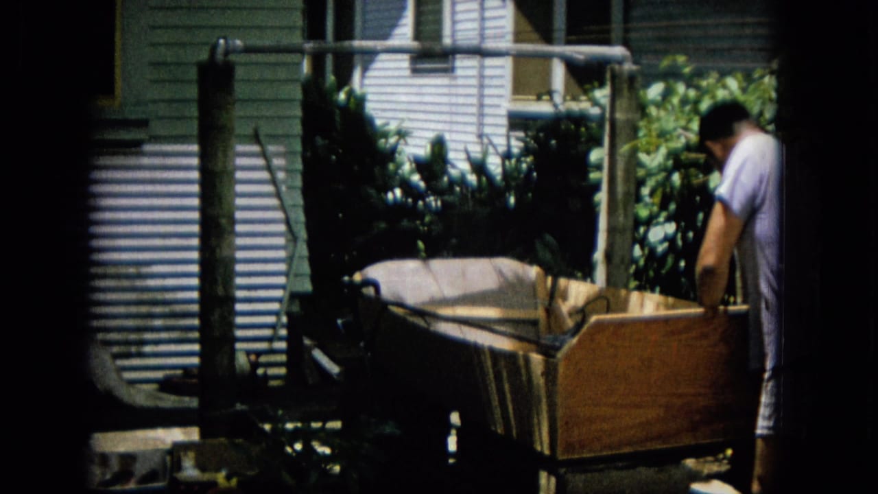 In archival 8mm home-movie footage from the supplied 1965 Hawaii metadata, a man stands partly turned away while working near a large wooden box-like structure in a leafy backyard.