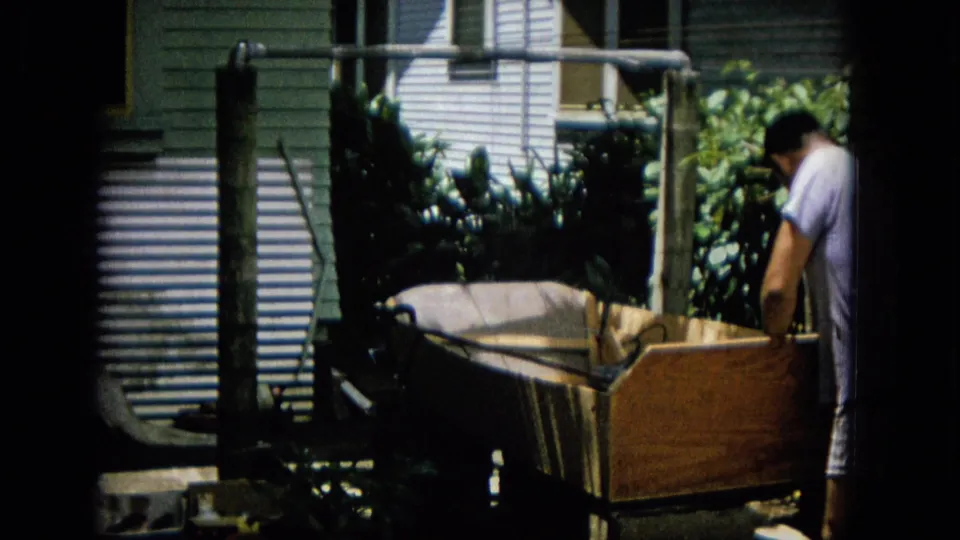 In archival 8mm home-movie footage from the supplied 1965 Hawaii metadata, a man stands partly turned away while working near a large wooden box-like structure in a leafy backyard.