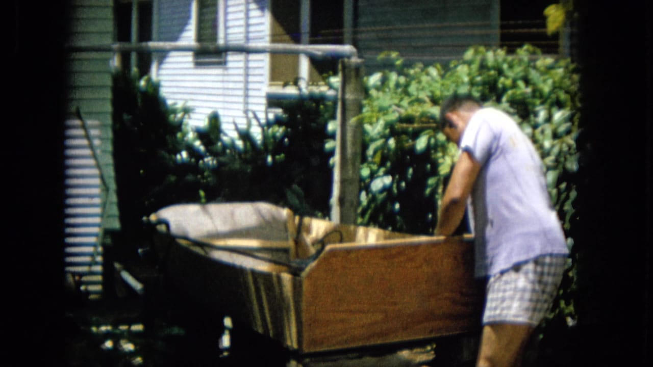 Man bends over a wooden structure near dense bushes and a white-sided house.