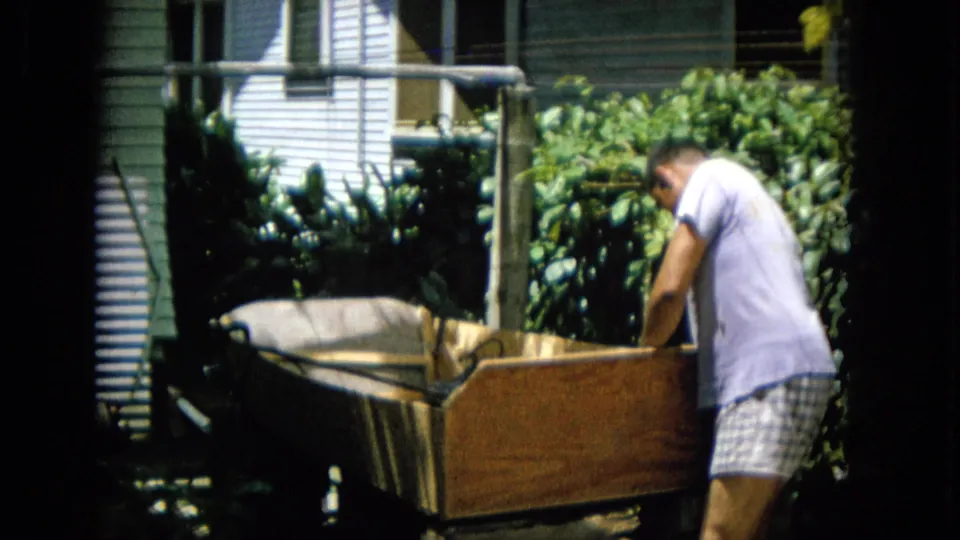 The man leans over the end of a large wooden structure in a sunny backyard, with dense foliage, siding, and a horizontal line or rail visible behind him.