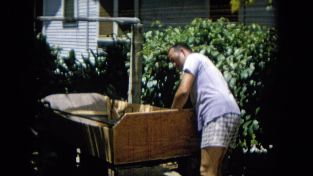 A man in a light shirt and plaid shorts bends close to the side of the wooden structure, framed by backyard greenery and partial house exterior details.