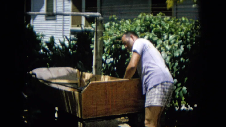 A man in a light shirt and plaid shorts bends close to the side of the wooden structure, framed by backyard greenery and partial house exterior details.