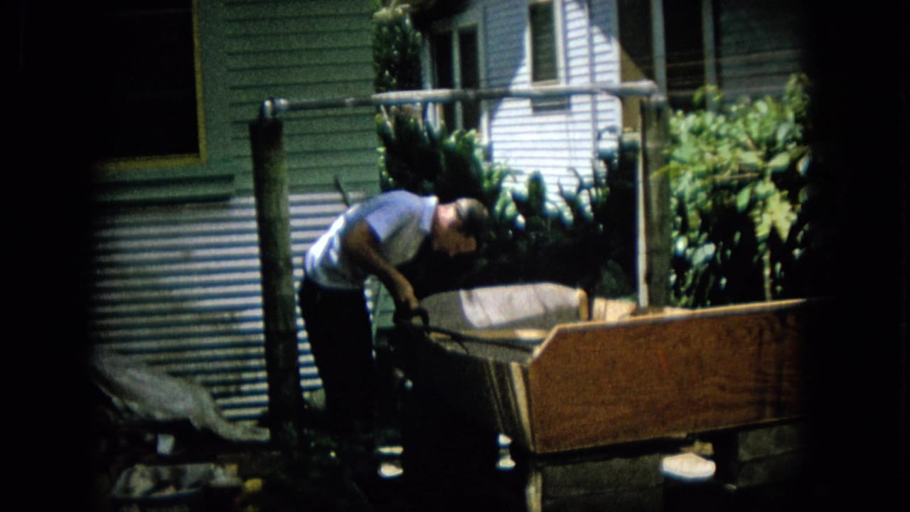 An adult man bends over the end of the wooden boat form near a light-colored building wall, posts, and leafy plants in the yard.