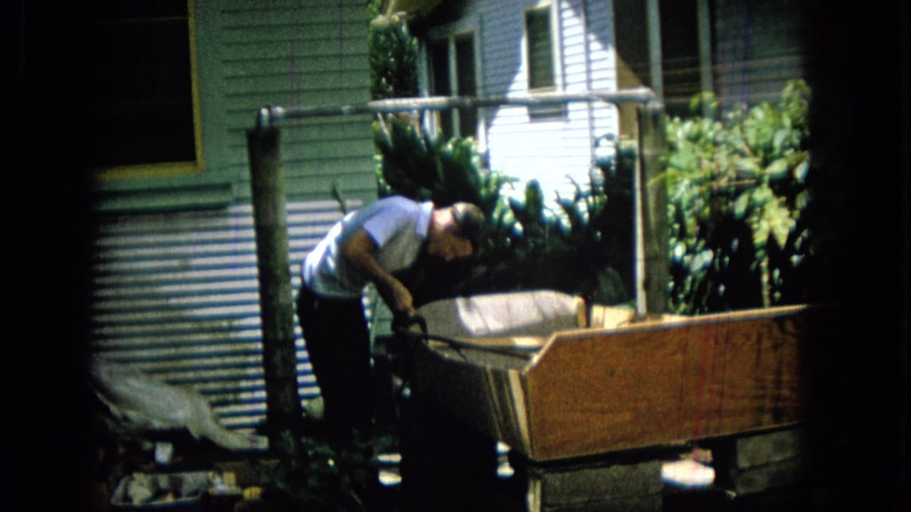 In 1965 Hawaii home-movie footage, a man in a light short-sleeve shirt bends over a wooden outdoor structure near house siding, windows, and leafy plants.