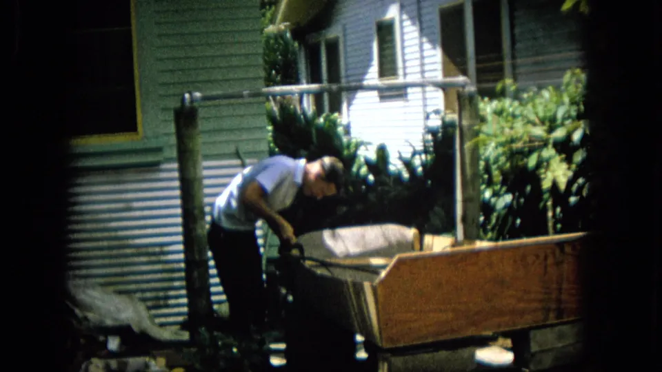 A man continues working over a wooden structure in a residential outdoor area, with light siding, a window, metal sheeting, and dense plants behind him.
