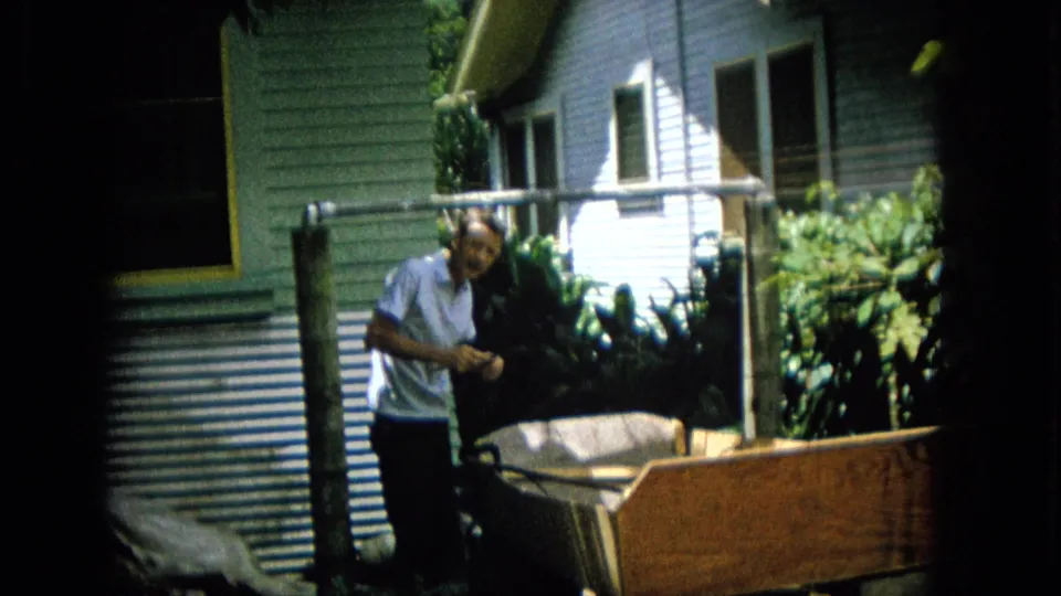 The man stands more upright beside the wooden outdoor structure, holding or inspecting something near a house wall and tropical-looking plants.