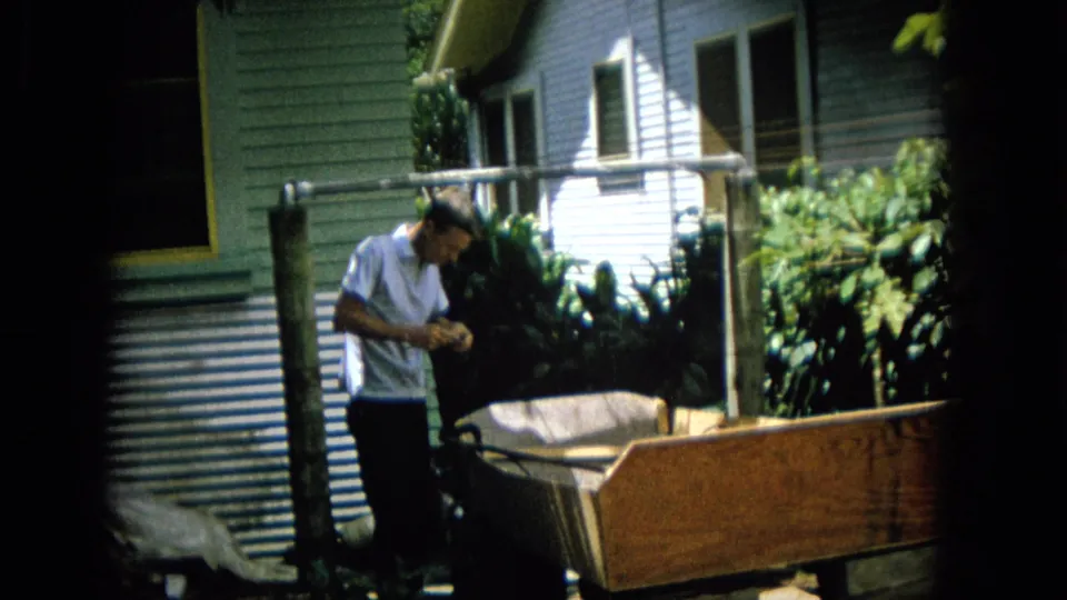 A man in a light shirt looks down at his hands beside a wooden structure, with a residential wall, window, pipes, and greenery visible in the background.