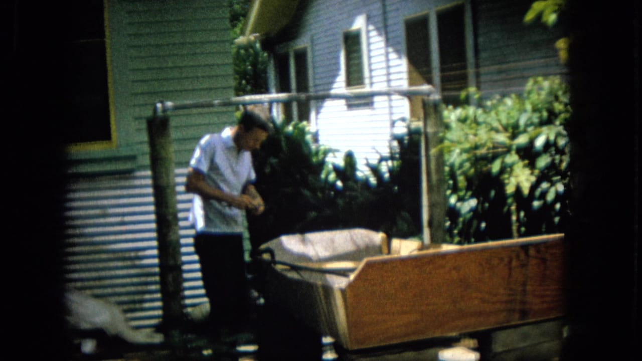 A man in a light short-sleeve shirt stands beside a large wooden box-like structure in a residential backyard, with house siding, pipe framing, and tropical foliage visible.