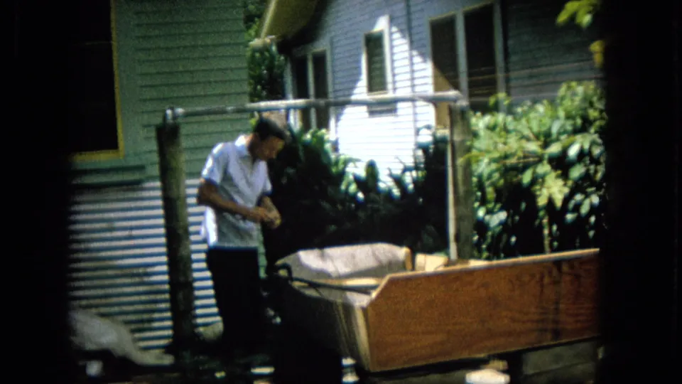 A man in a light short-sleeve shirt stands beside a large wooden box-like structure in a residential backyard, with house siding, pipe framing, and tropical foliage visible.