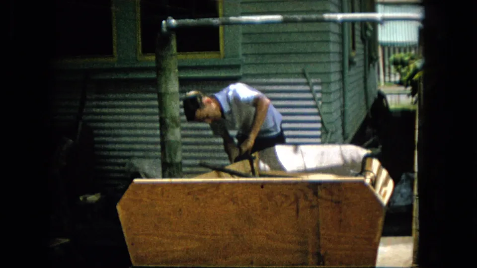 A man bends forward over the wooden structure and grips a dark tool or handle, with corrugated siding, a window, and yard foliage in the background.