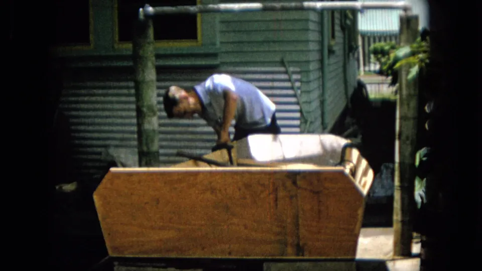 The man remains bent over the wooden structure, holding a dark handle or tool, with house siding, a pipe frame, and bright foliage behind the work area.