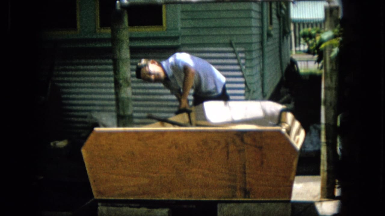 In 1965 archival 8mm footage from the United States, a man in a light shirt leans over a large wooden project beside green exterior siding and a corrugated metal surface.