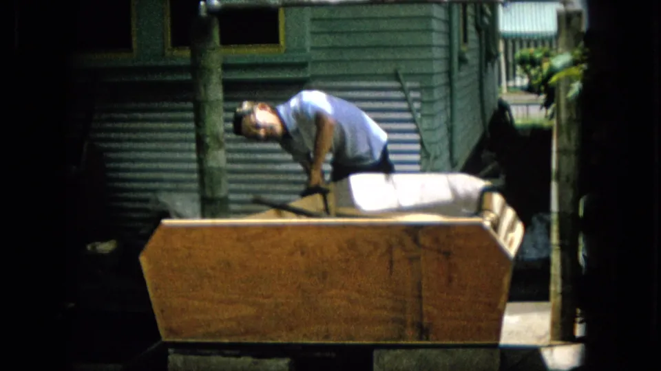 In 1965 archival 8mm footage from the United States, a man in a light shirt leans over a large wooden project beside green exterior siding and a corrugated metal surface.