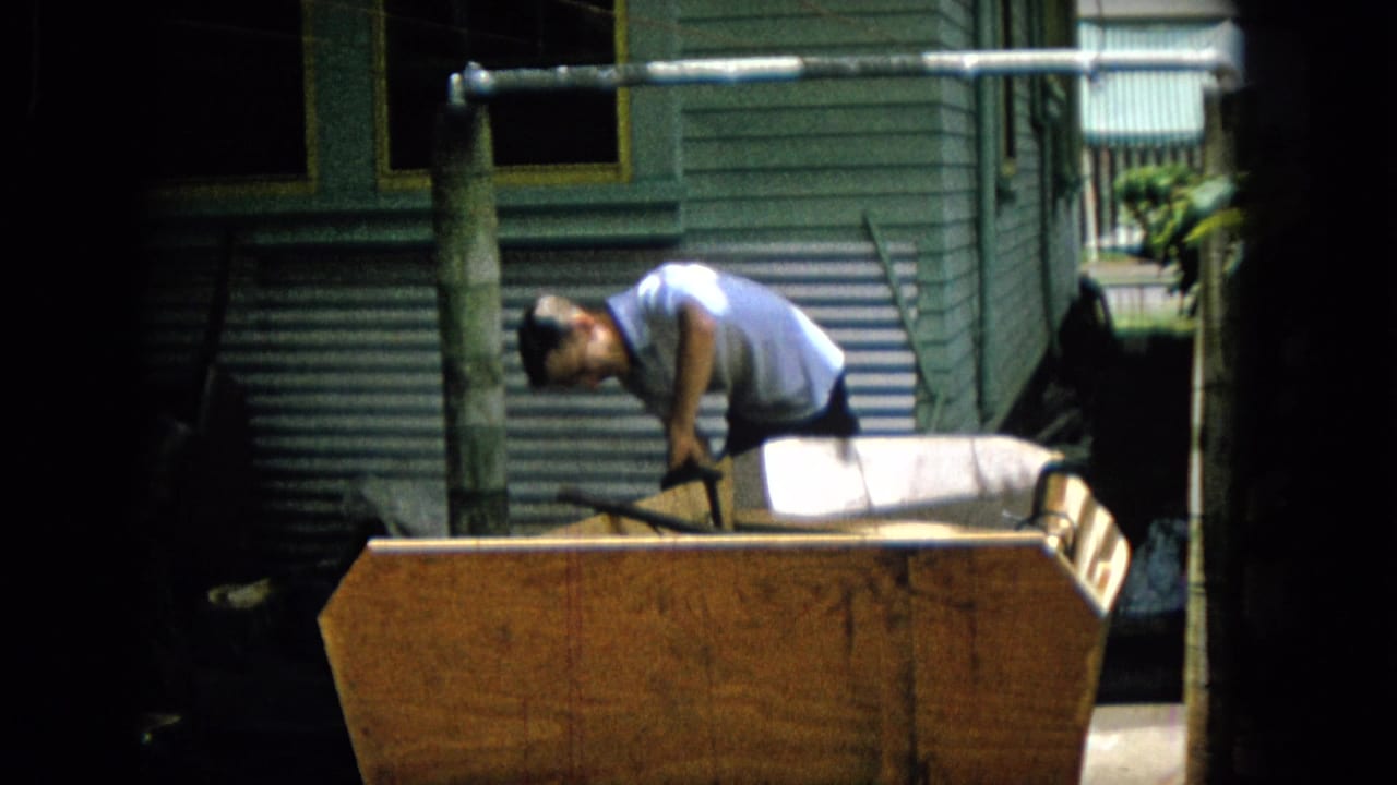 The man remains bent over the wooden object, with a light cushion or padded surface and exterior wall details visible behind him in the home-movie frame.