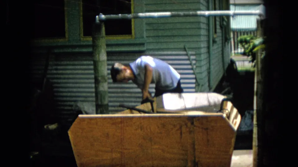 The man remains bent over the wooden object, with a light cushion or padded surface and exterior wall details visible behind him in the home-movie frame.