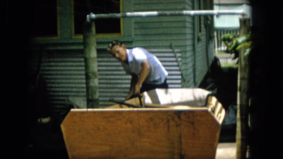 The man briefly looks toward the camera while standing over the large wooden object, making his face more visible in the 1965 home-movie footage.