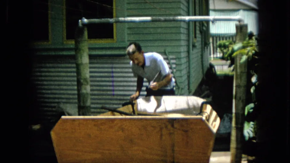 Near the end of the clip, the man stands behind the large wooden project with a tool in hand, framed by residential siding, posts, and strong sunlight.