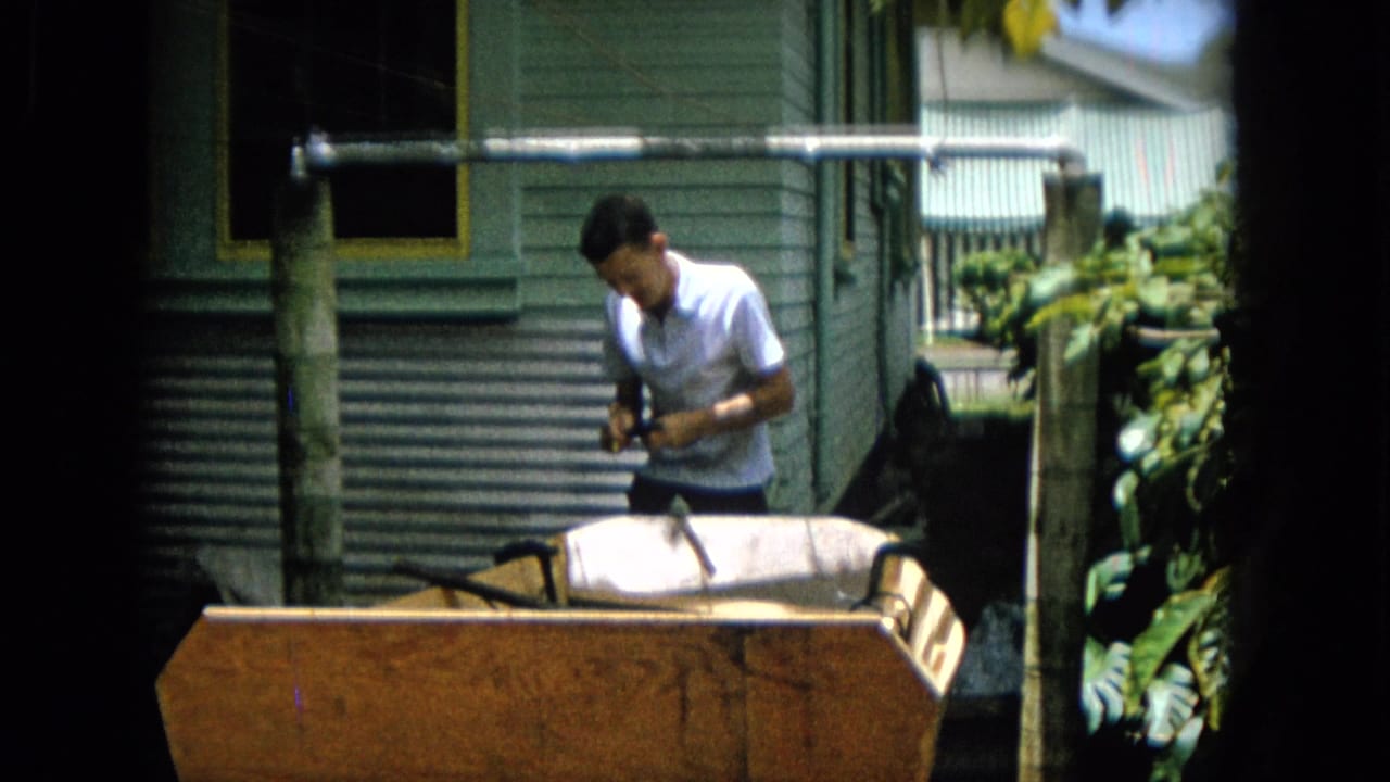 Man in a white shirt stands beside a wooden boat hull outside a green-sided house.