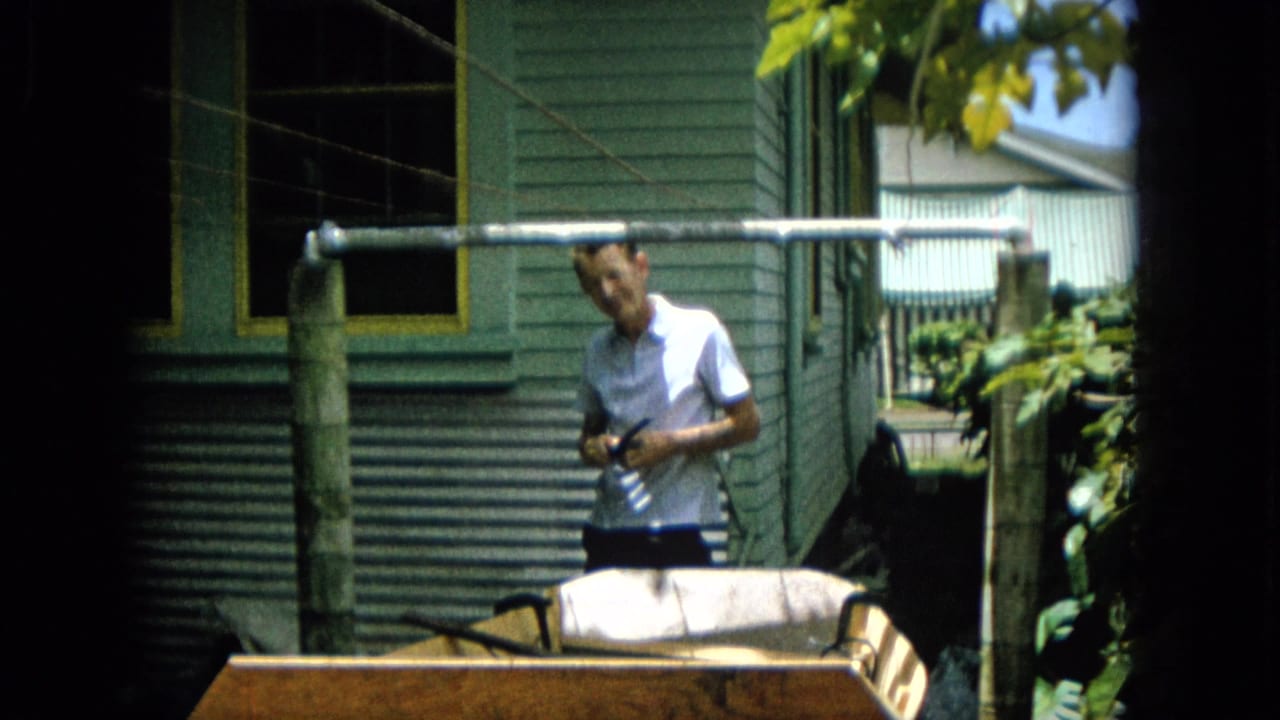A man in a white shirt looks toward the camera from behind a homemade wooden boat hull outside a green-sided residence in 1965 Hawaii footage.
