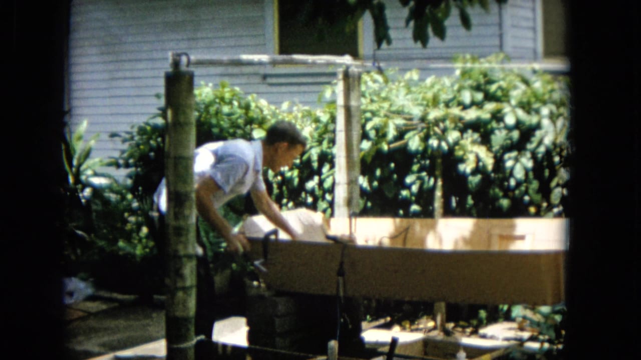 Man bends over the wooden boat hull near sunlit shrubs and yard posts.