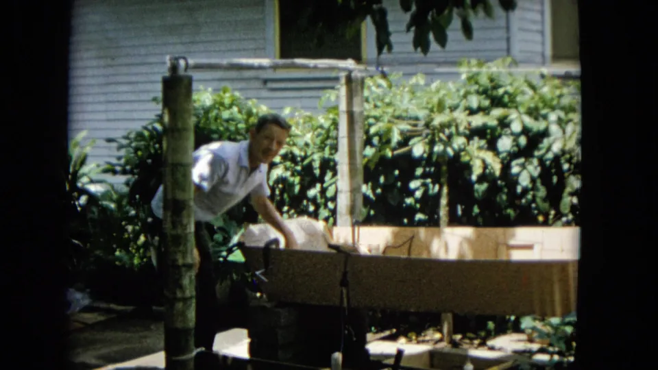 A man works beside the homemade wooden boat hull in a sunny residential yard, partly turning toward the camera in the 1965 Hawaii clip.