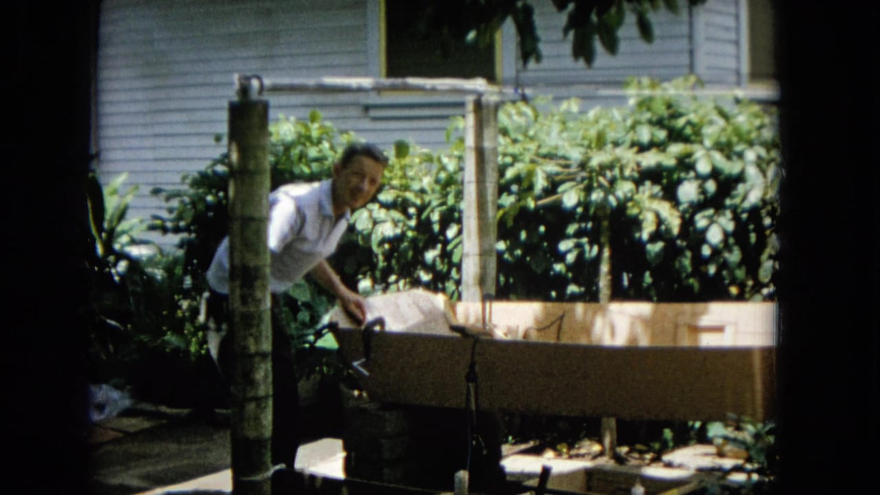 In 1965 Hawaii home-movie footage, an adult man in a white short-sleeved shirt leans over a long wooden structure set among garden foliage beside a light-sided house.