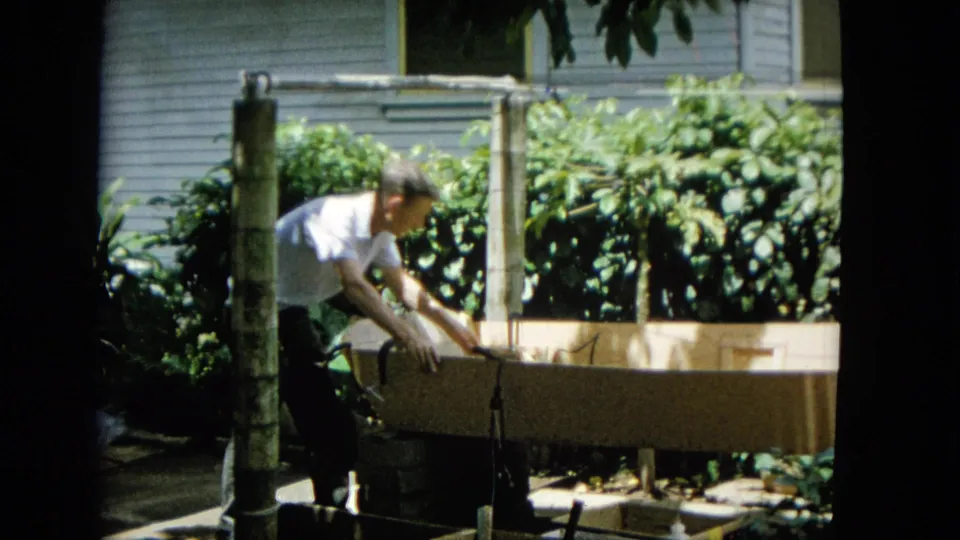 The man works with a hand-held power tool over the wooden frame, with bamboo-like supports, dense plants, and house siding visible behind him.