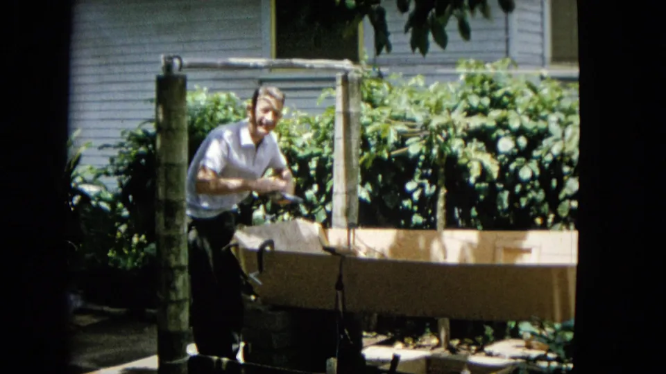 The adult man stands beside the partially built wooden structure in the tropical yard, briefly facing the camera with foliage and a light-sided house in the background.