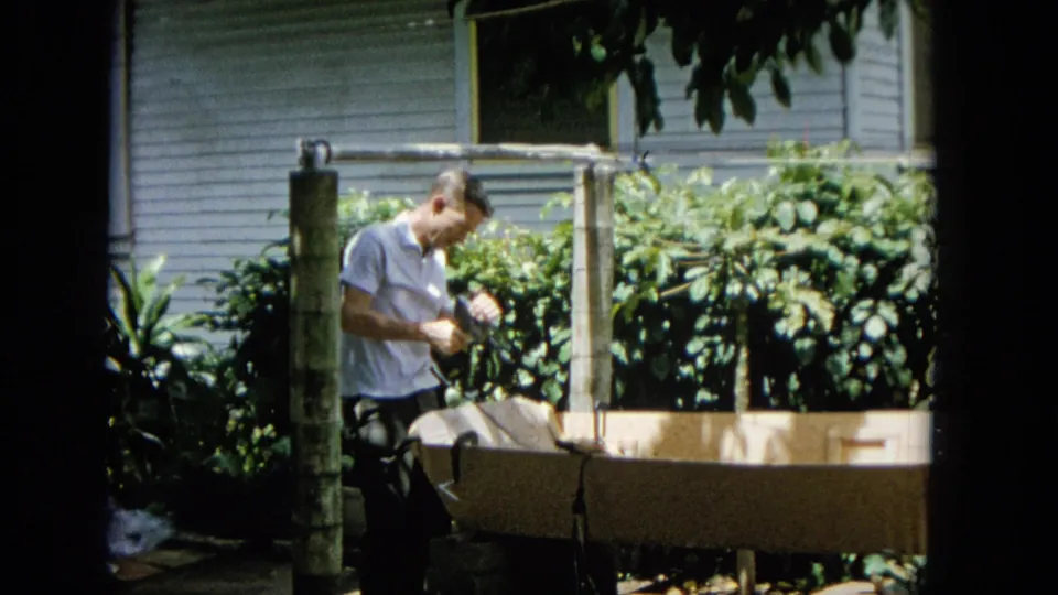 The man looks down at a hand-held tool or drill beside the long wooden structure, framed by tropical plants and the exterior wall of a house.