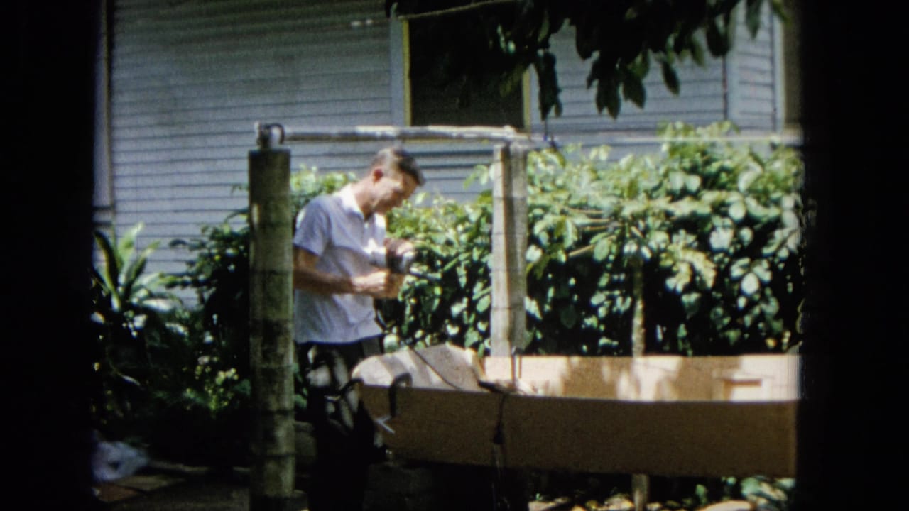 In this 1965 United States home-movie frame, an adult man in a white short-sleeve shirt appears to tend a wooden garden frame amid dense tropical foliage and a nearby house exterior.