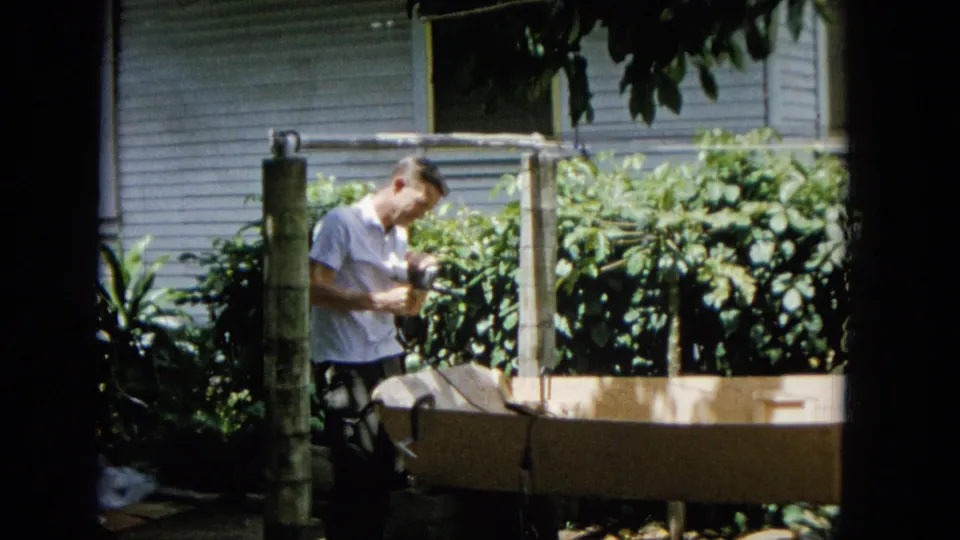 In this 1965 United States home-movie frame, an adult man in a white short-sleeve shirt appears to tend a wooden garden frame amid dense tropical foliage and a nearby house exterior.
