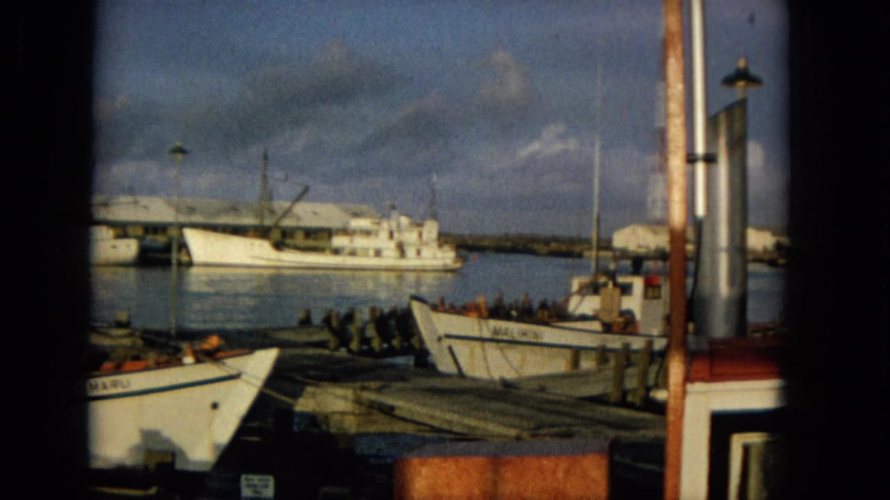 1965 Hawaii harbor view on 8mm film, with small boats at wooden docks, a larger vessel in the distance, calm water, and shoreline buildings under a partly cloudy sky.