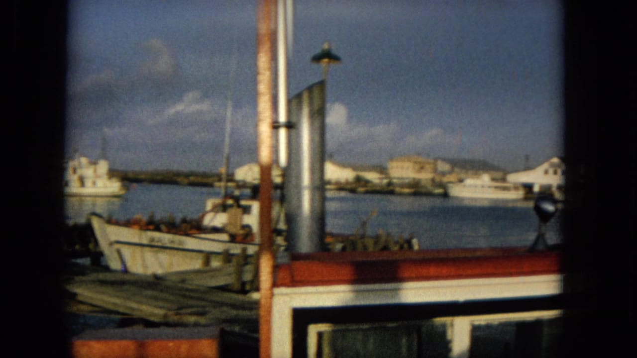 1965 Hawaii harbor footage showing moored small boats, dock timbers, a red-and-white boat cabin edge, shoreline buildings, and calm blue water.