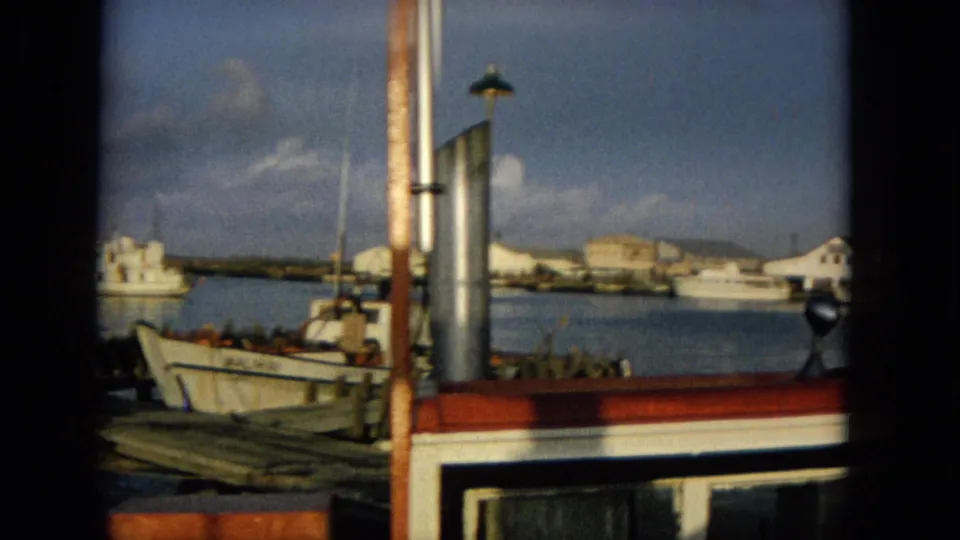 1965 Hawaii harbor footage showing moored small boats, dock timbers, a red-and-white boat cabin edge, shoreline buildings, and calm blue water.