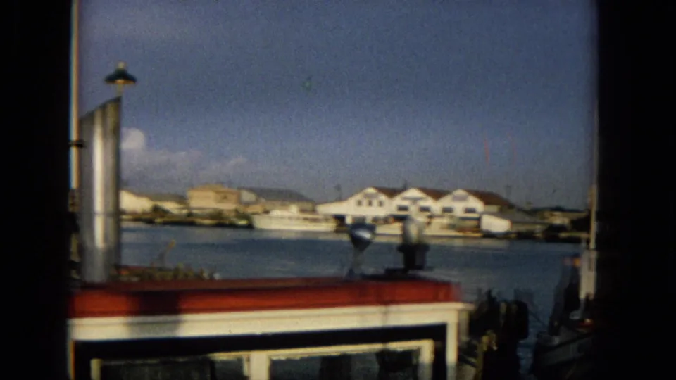1965 Hawaii harbor scene with a boat cabin foreground, a small vessel across the water, white waterfront buildings, distant hills, and partly cloudy sky.