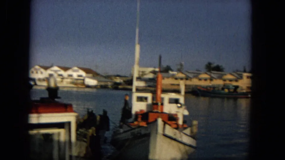 1965 Hawaii harbor view with a small white boat near a wooden dock, distant people on the pier, waterfront buildings, calm water, and boat masts in the background.