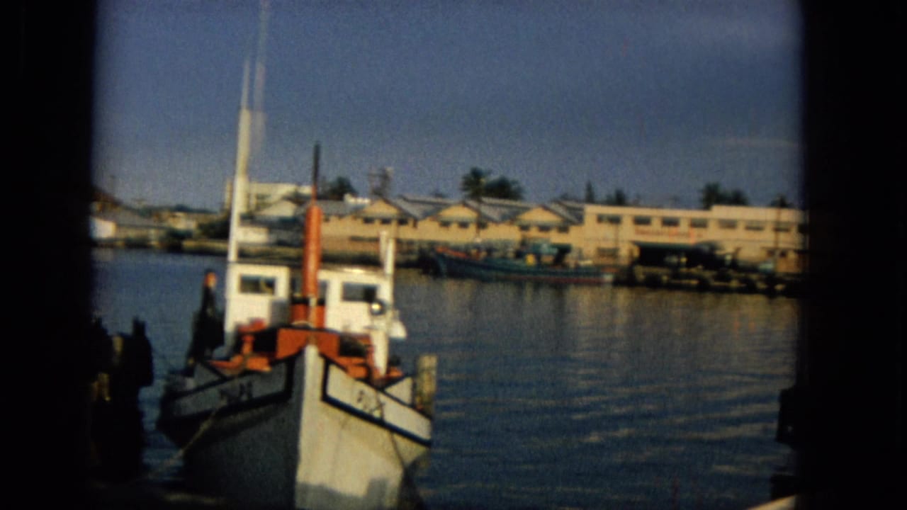 1965 Hawaii 8mm frame showing a small boat near dock pilings, calm harbor water, and low shoreline buildings in soft late-day light.