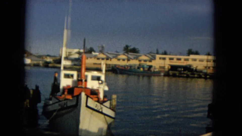 1965 Hawaii 8mm frame showing a small boat near dock pilings, calm harbor water, and low shoreline buildings in soft late-day light.