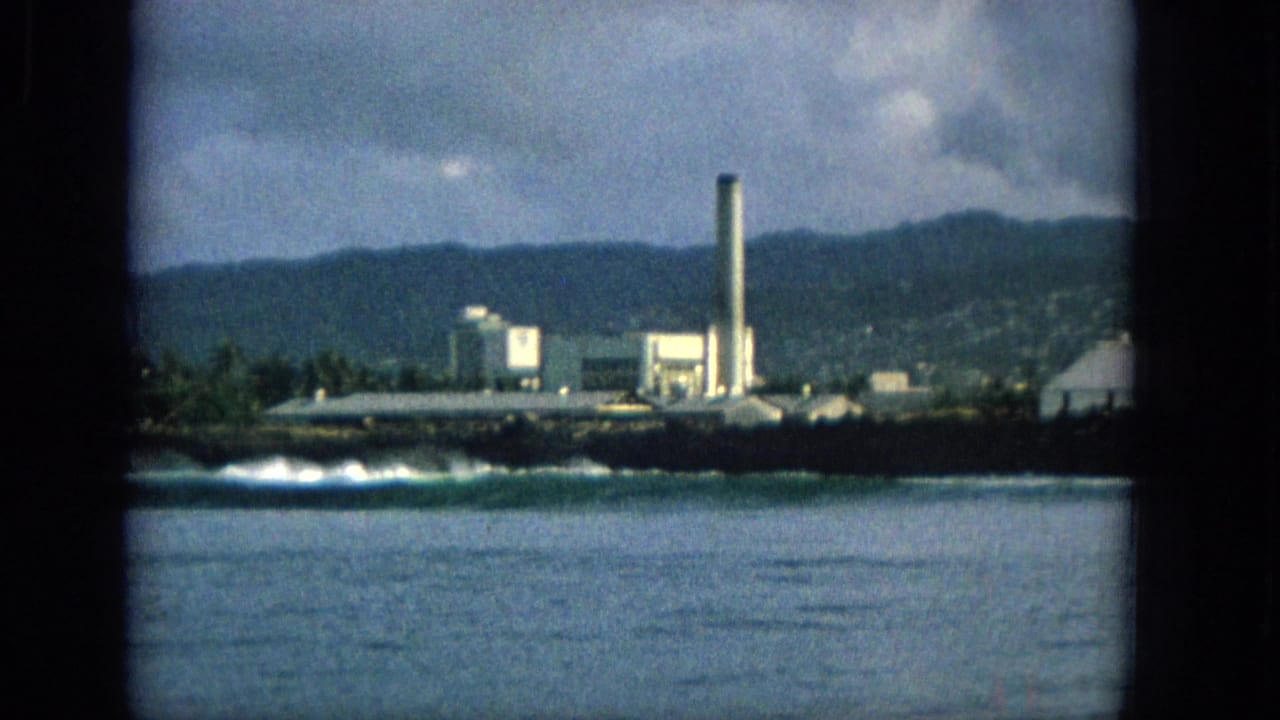 1965 Hawaii 8mm frame showing a distant coastal industrial site with a tall smokestack, surf, dark shoreline, mountains, and clouds.
