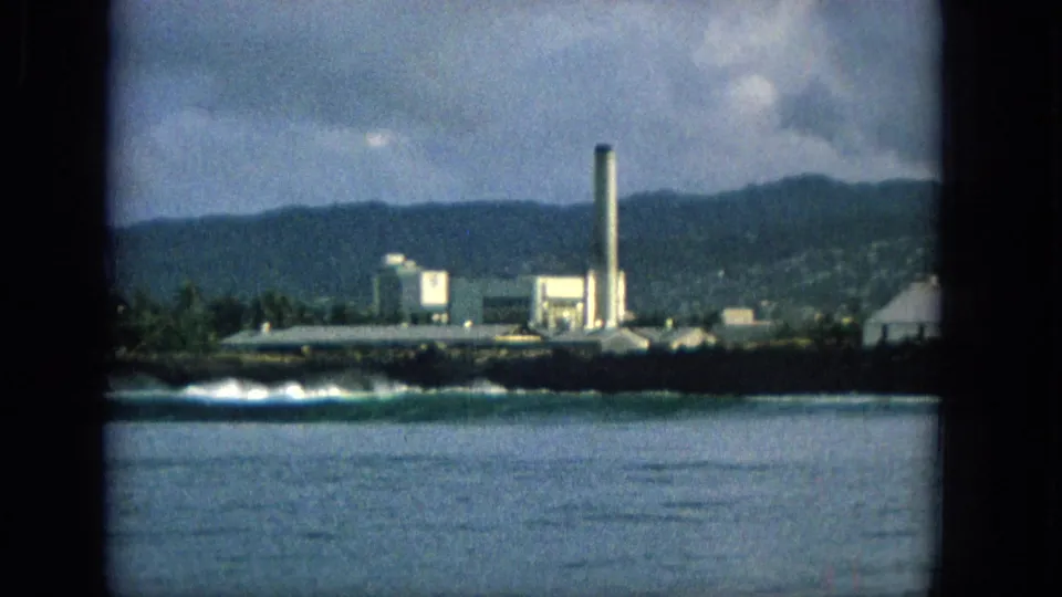 1965 Hawaii 8mm frame showing a distant coastal industrial site with a tall smokestack, surf, dark shoreline, mountains, and clouds.