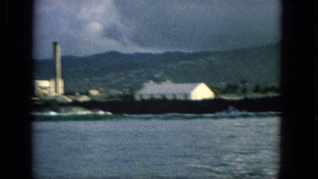1965 Hawaii 8mm frame showing a white building near the shoreline, breaking surf, a distant smokestack, mountains, and overcast sky.