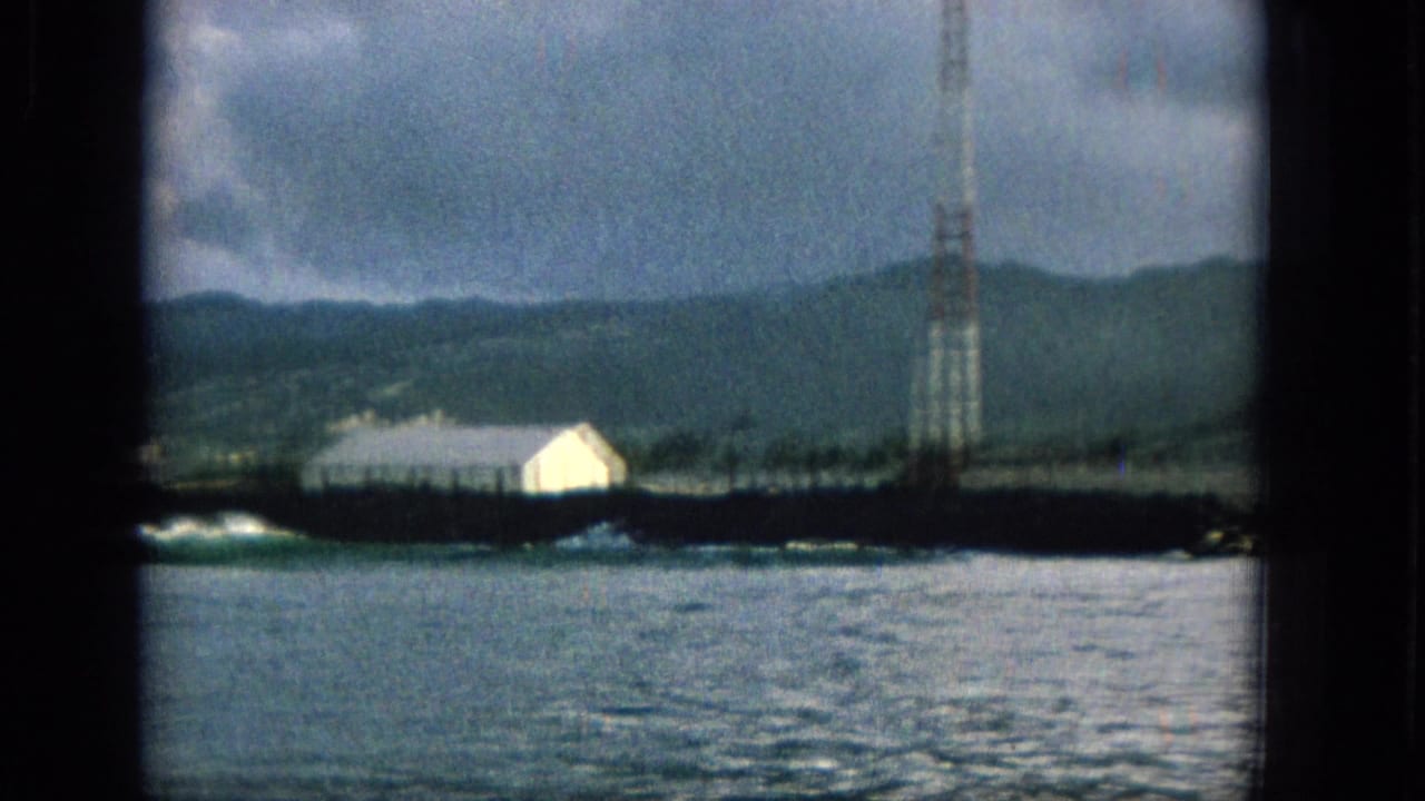 1965 Hawaii 8mm frame showing calm coastal water, a low shoreline, a white building, distant green hills, and a tall communications-style tower under cloudy sky.
