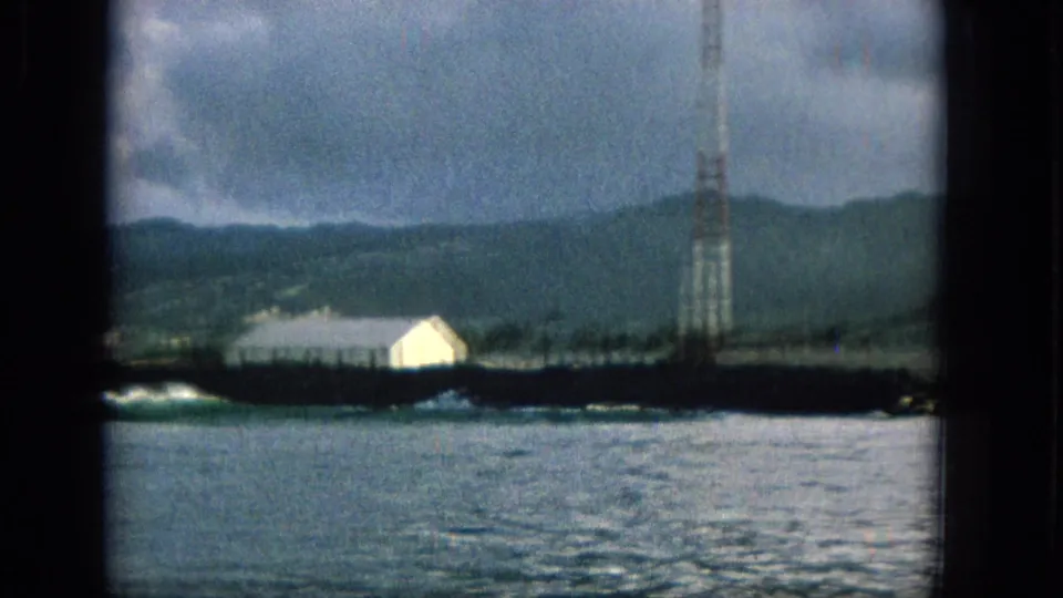 1965 Hawaii 8mm frame showing calm coastal water, a low shoreline, a white building, distant green hills, and a tall communications-style tower under cloudy sky.