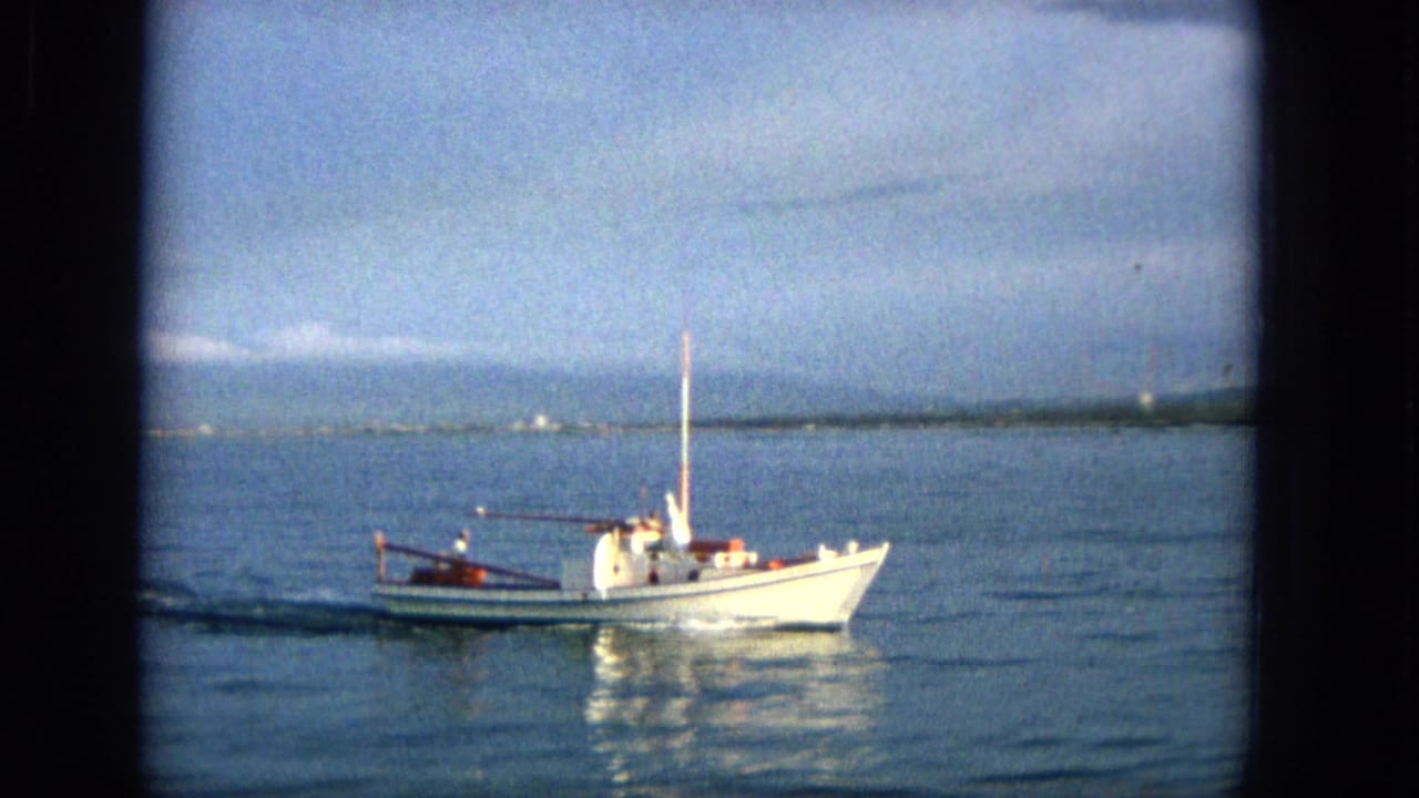 1965 Hawaii 8mm frame of a small white motorboat with red trim and a tall mast moving across calm blue water with distant hills and shoreline behind it.