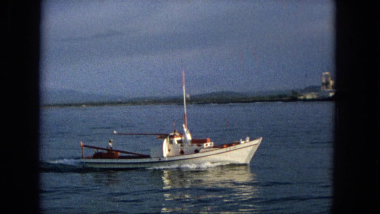 1965 Hawaii 8mm frame showing the white motorboat closer in profile, with a small wake, reflective water, distant shoreline, and hazy hills beyond.