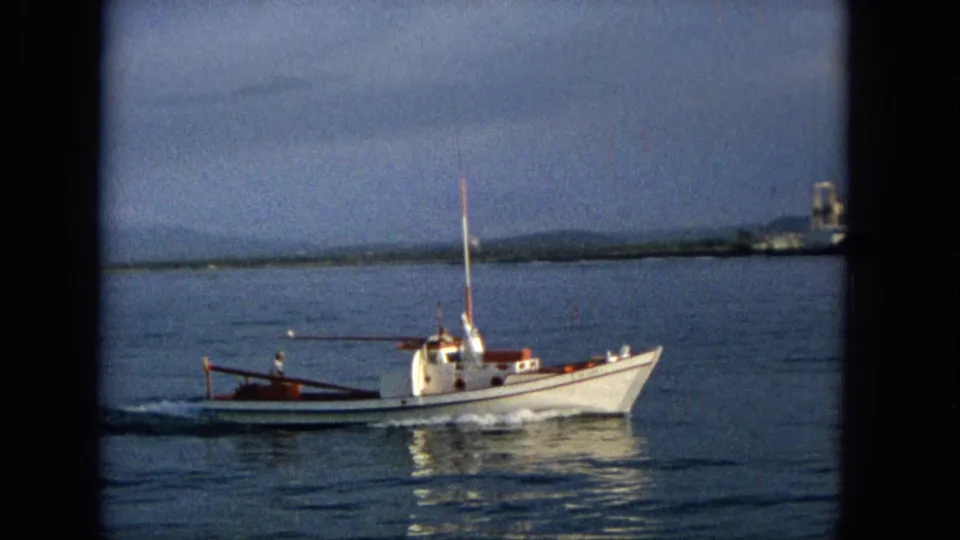 1965 Hawaii 8mm frame showing the white motorboat closer in profile, with a small wake, reflective water, distant shoreline, and hazy hills beyond.