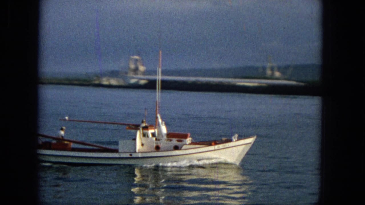 1965 Hawaii 8mm frame with a closer side view of the white motorboat, red cabin trim, mast, rippling reflection, low shoreline, and distant hills.