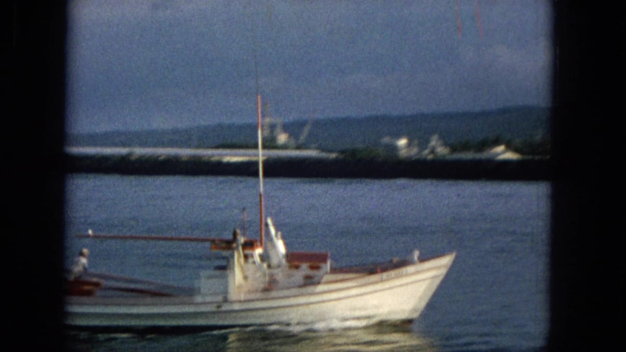 A small white boat with red trim passes across calm water near a distant Hawaiian shoreline in 1965 home-movie footage.